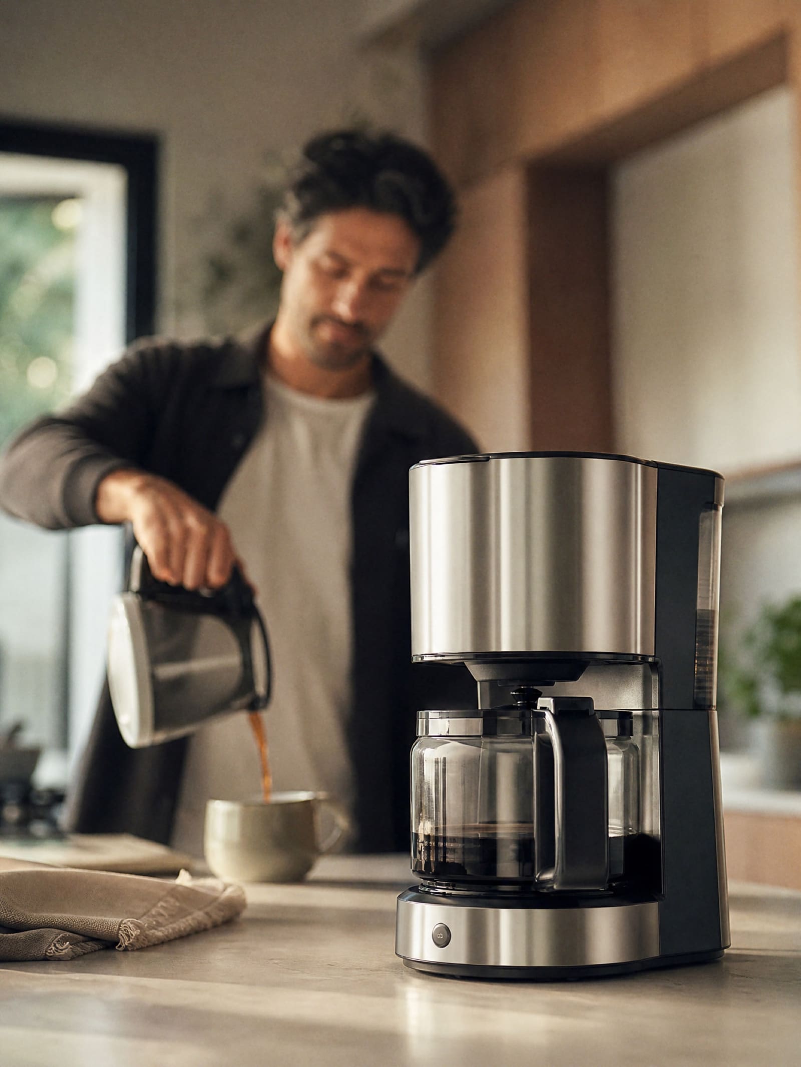 Hombre que sirve café en su cocina con la cafetera nítida en primer término