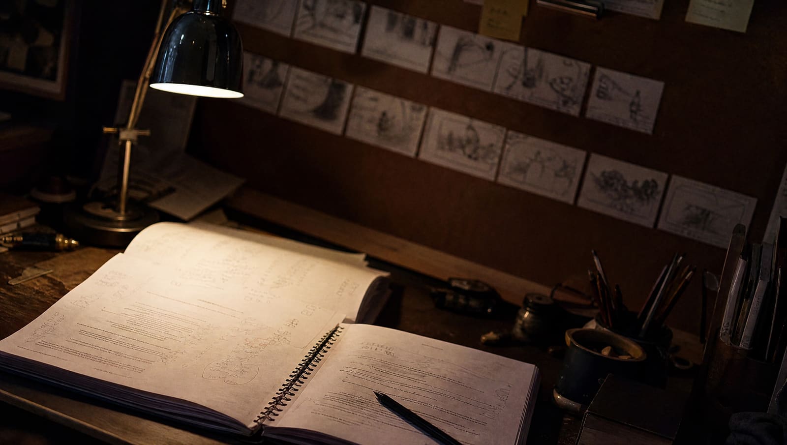 Mesa de trabajo de noche con un guion impreso abierto, un lápiz encima y una fila de viñetas de storyboard dibujadas a lápiz clavadas en un corcho detrás