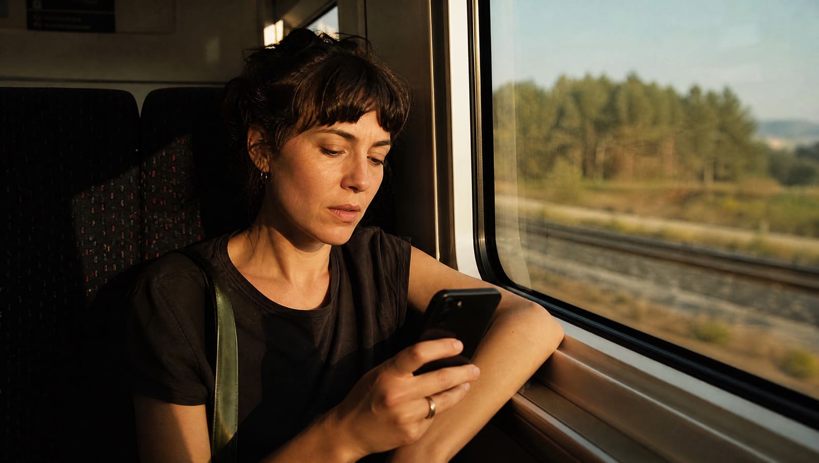 Una mujer adulta en el asiento de un tren desliza con el pulgar un feed de vídeos verticales en su móvil, con luz de tarde por la ventanilla y la pantalla ligeramente desenfocada
