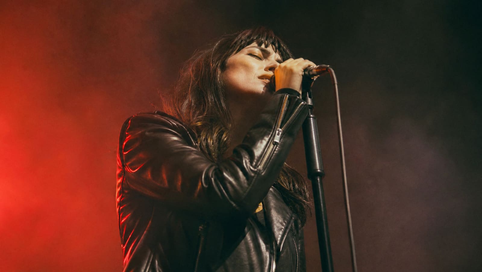 Una cantante adulta con chaqueta de cuero canta con los ojos cerrados en un escenario vacío, envuelta en humo y en una luz roja que se funde con un paisaje irreal al fondo